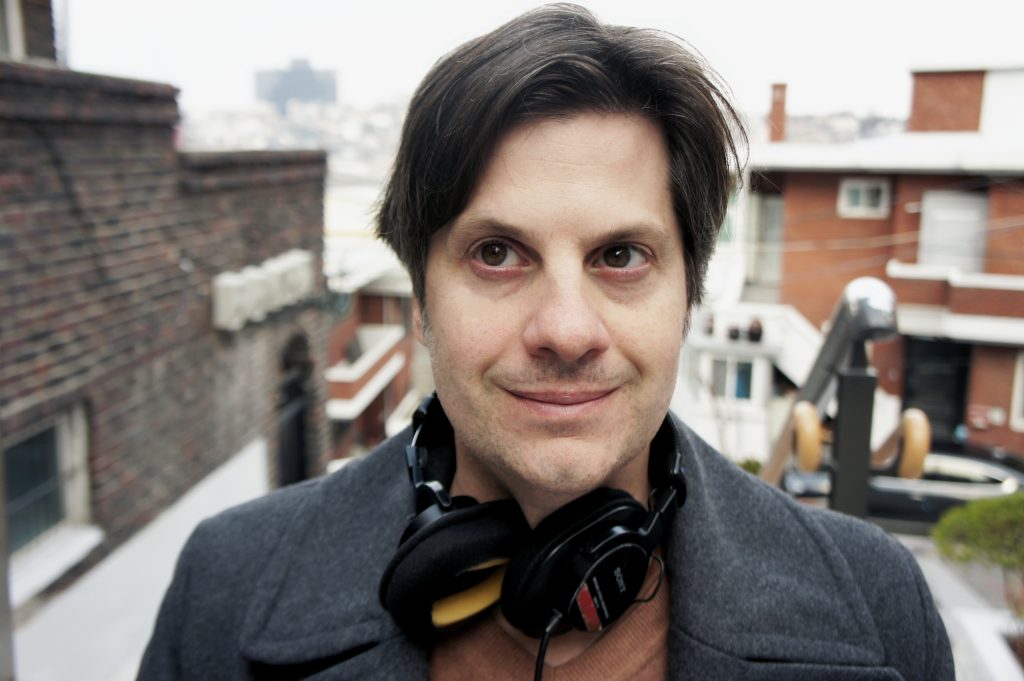 Jason Strother, a light skinned mail with dark hair, is pictured with a pair of headphones wrapped around his neck in front of an urban backdrop. 